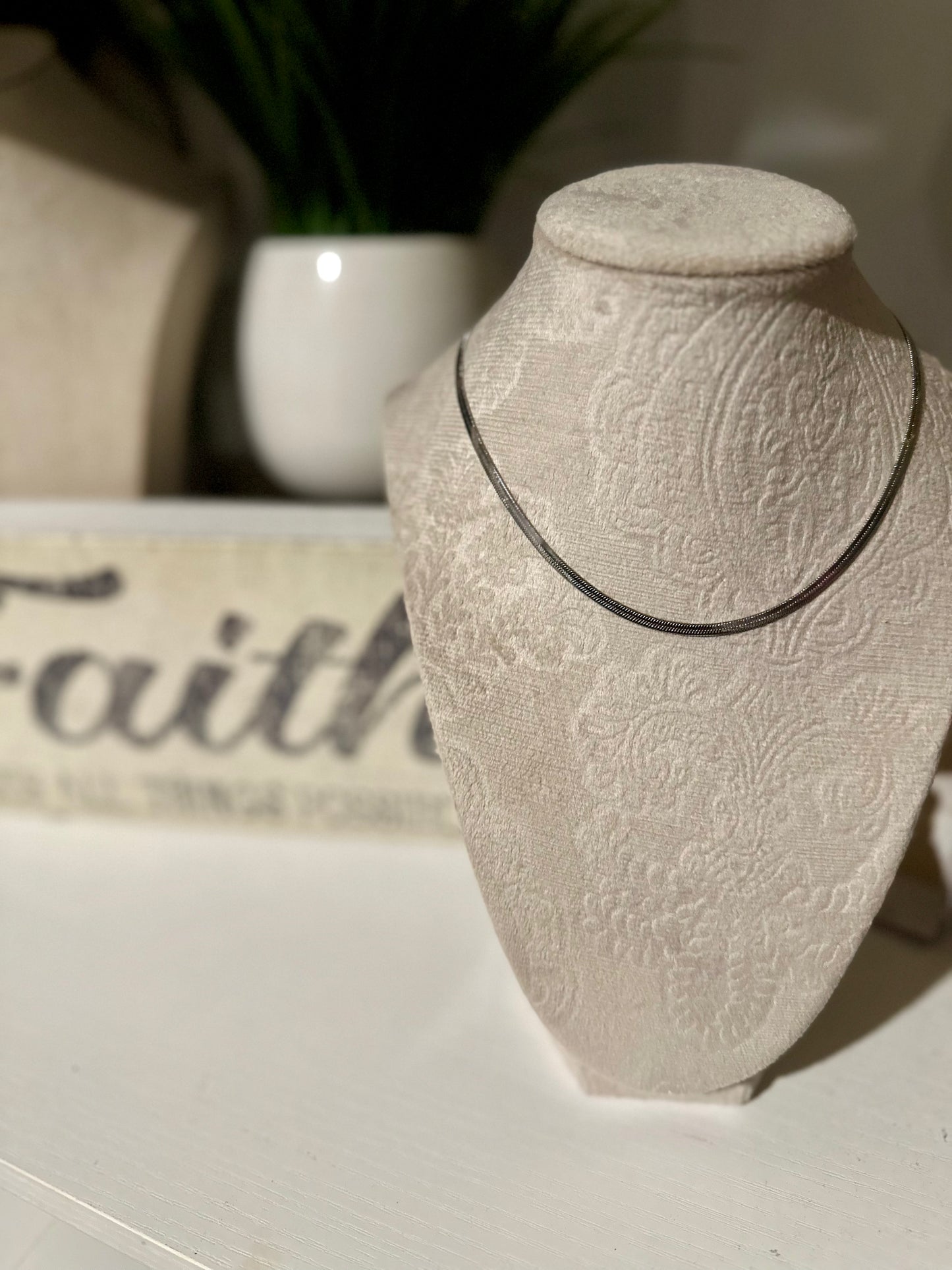 Necklace displayed on a textured stone bust with a blurred 'Faith' sign in the background.